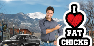 A man points towards a "I heart fat chicks" sign with a black 2019 Chevy Impala and mountains in back