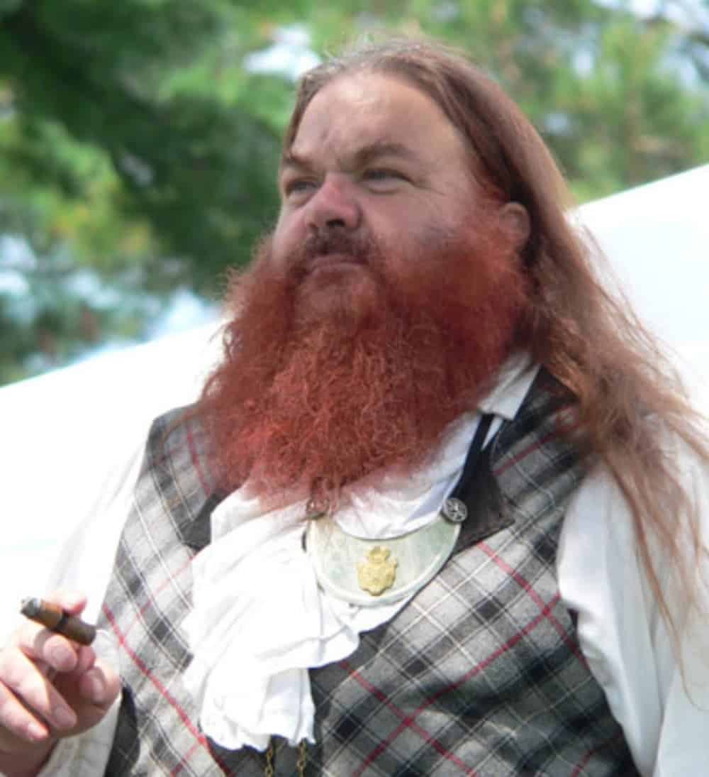 A Scottish man in plaid with a red beard and long red hair is holding a cigar.