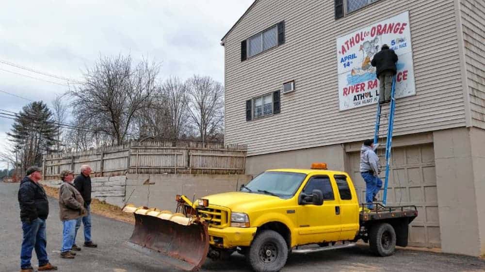 Two men are standing on the back of a flat bed pickup truck with an unsafe ladder while other men watch.