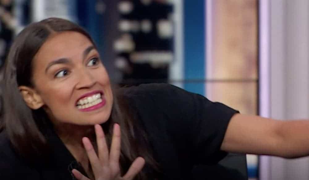 In a still image from an interview, Alexandria Ocasio-Cortez is shown with a wide eyed smile and her hand up.