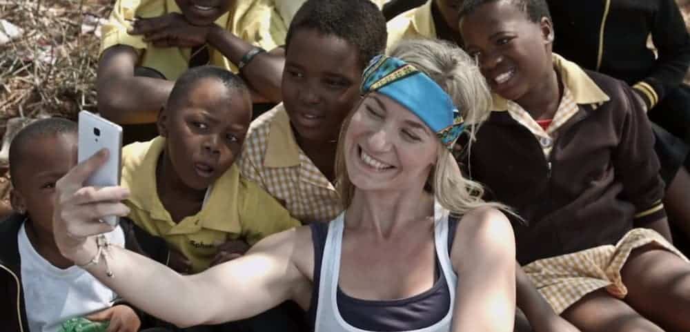 A white woman is taking a selfie with a group of presumably African kids.