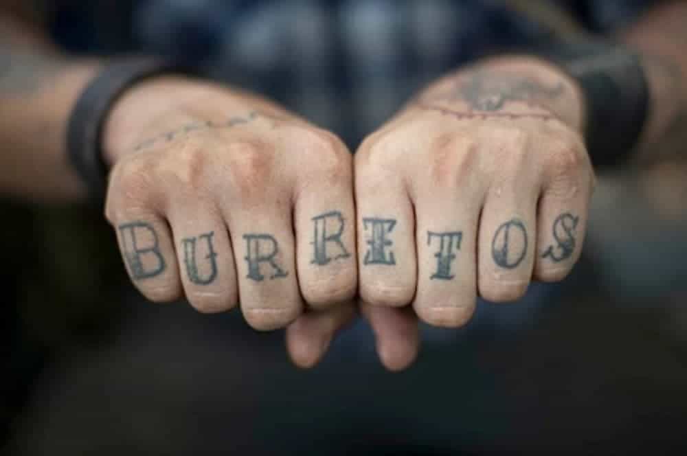 A tattoo across a man's knuckles reads 'burritos'.