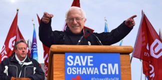 Bernie Sanders is shown behind a podium with a Save Oshawa GM on the sign as part of live auto news.