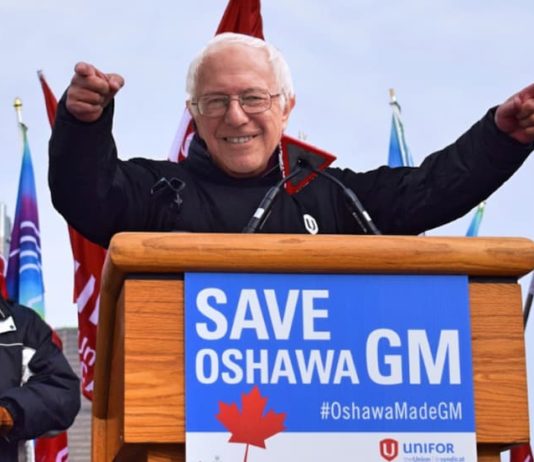 Bernie Sanders is shown behind a podium with a Save Oshawa GM on the sign as part of live auto news.