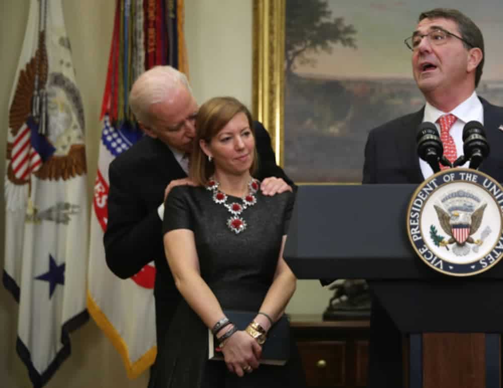 Joe Biden is smelling an uncomfortable woman from behind at a political event.