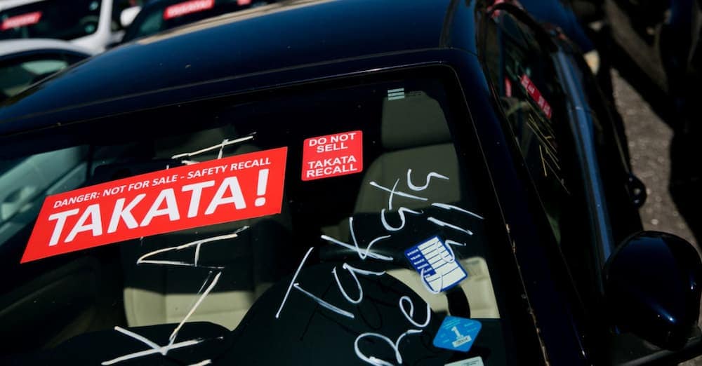 A car window is shown with Takata recall stickers to prevent sale as they are being recalled in live auto news.