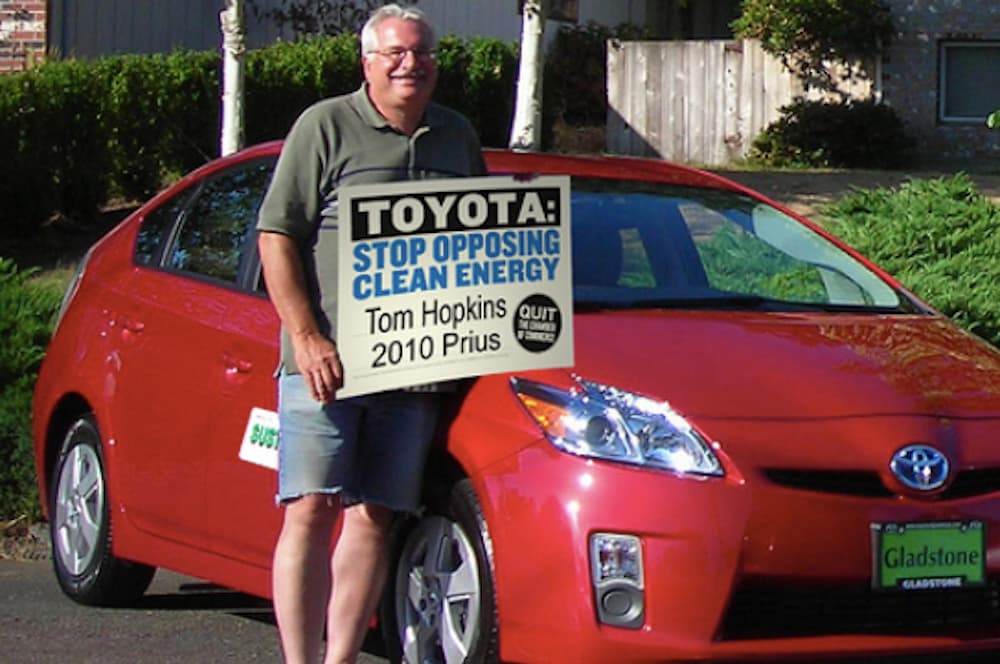 A man is holding a clean energy sign in front of his red Prius.