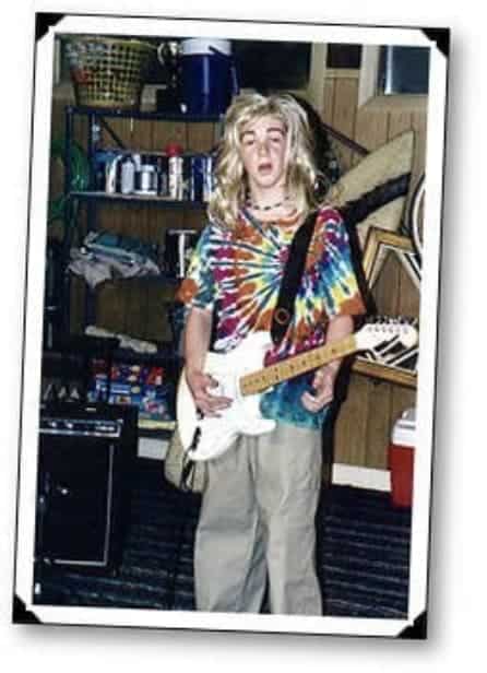 Drake Bell is dressed up with a tie dye shirt and long blond wig while holding a guitar.