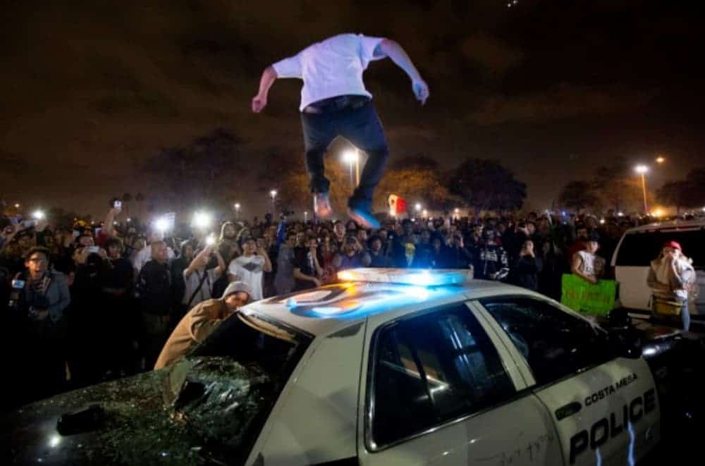A riot is happening while someone jumps on a police car.