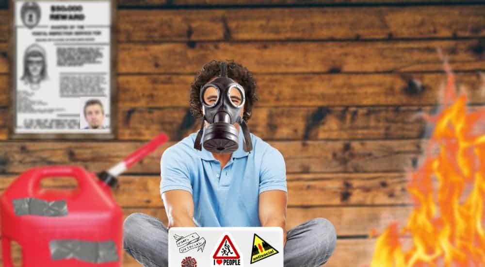 A man in a gas mask is next to a gas can and a fire while looking at live auto news on his laptop.