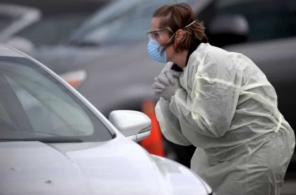A woman is leaning towards a car while dressed in gear to prevent Coronavirus, which is an issue in live auto news.