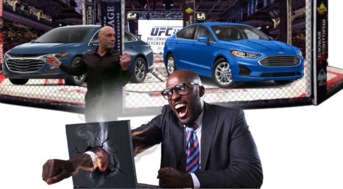 A man is punching through a computer in front of Joe Rogan and a UFC octagon showing a fight between a blue 2020 Chevy Malibu vs 2020 Ford Fusion.