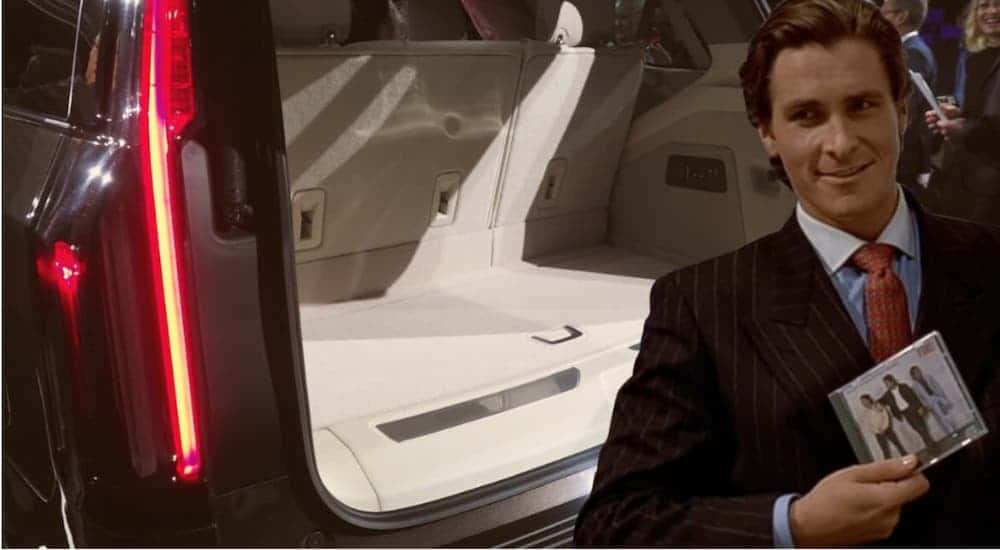 Patrick Bateman from American Psycho is holding a CD in front of a opened Cadillac SUV cargo space.