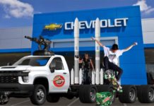 A skateboarder is doing a trick in front of a used Chevy truck fitted with rockets and gun turrets next to a box of mountain dew and a Terminator, in front of a Chevy dealership.