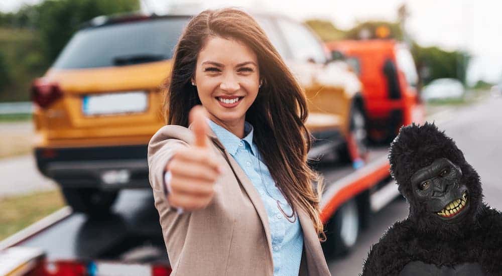 A woman has a thumbs up in front of a new car on a tow truck next to a gorilla suit.