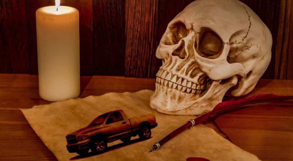 A skull, candle, quill and parchment with a Ram truck on it are shown on a table.