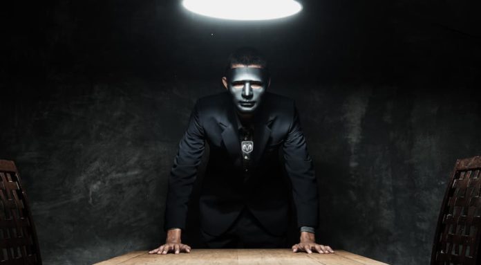 A man with a black mask and Ram logo is looking over a table in a dark room.