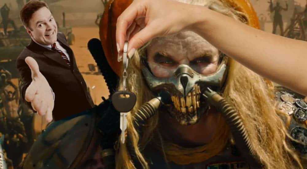 A character from Mad Max Fury Road is in front of a car salesman and being handed car keys.