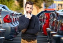 A man with his hand on his chin questioningly is standing between two sets of cars at a Chevy Dealer in Norfolk.