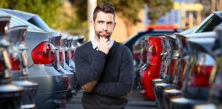 A man with his hand on his chin questioningly is standing between two sets of cars at a Chevy Dealer in Norfolk.