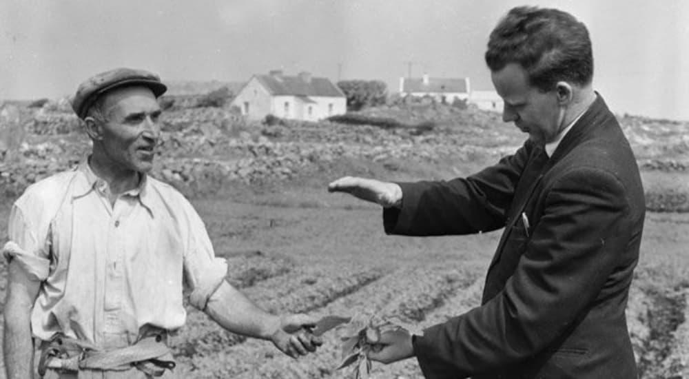 A black and white photo of a business man in a suit and a farmer are standing in a field exchanging leaves.