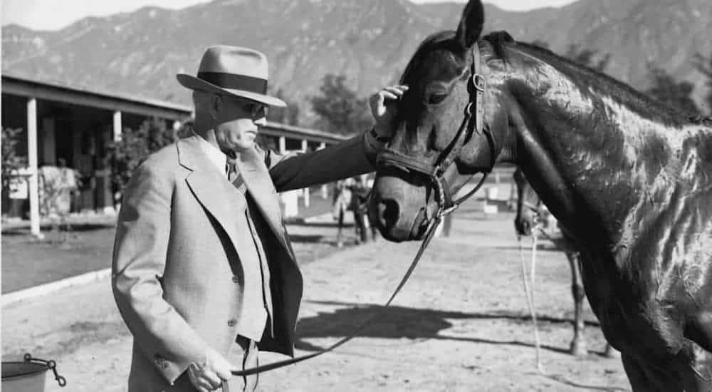 A black and white photo of an old man in a suit and hat who is petting a horse.