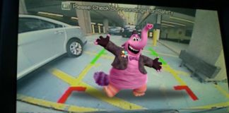 A pink elephant with a jacket and top hat is standing behind a car shown on the back up camera screen.