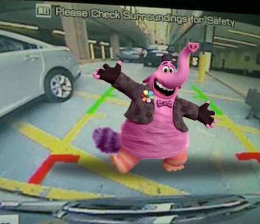 A pink elephant with a jacket and top hat is standing behind a car shown on the back up camera screen.