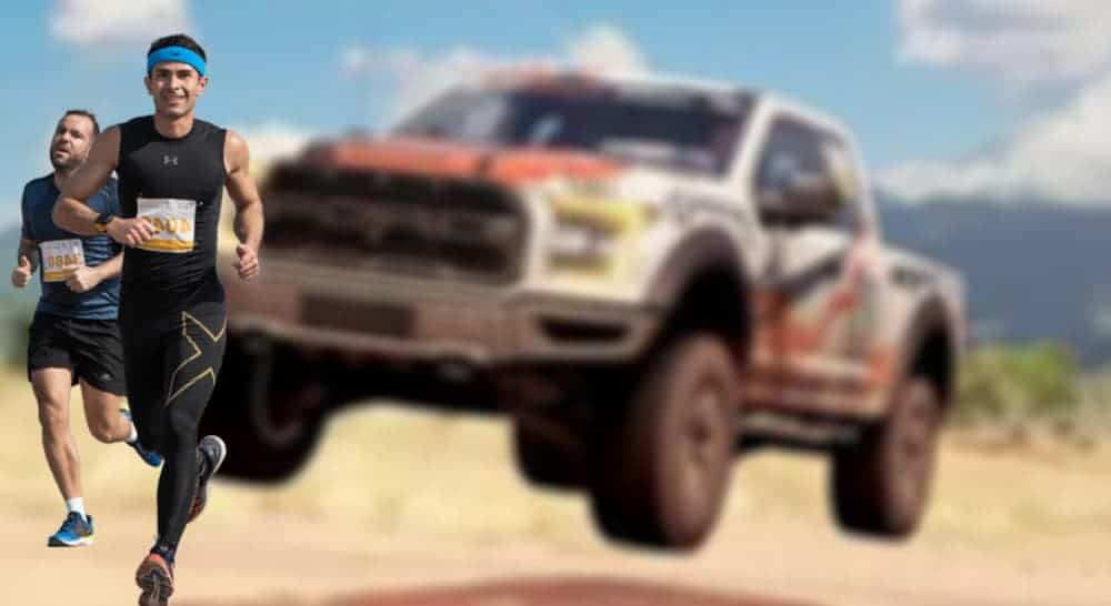 A Ford Raptor race truck is driving in the desert behind two runners.