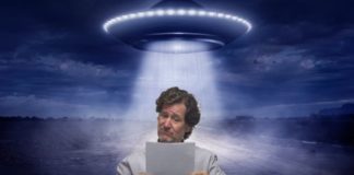 A man is reading a paper while a spaceship is beaming light over him.