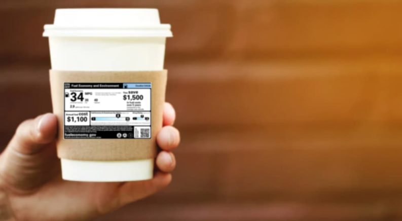 A hand is shown holding a cup of coffee with a vehicle sales window sticker on it.