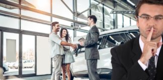 A salesman is making a silence gesture while a couple is shaking hands with a salesman in the background at a car dealer.