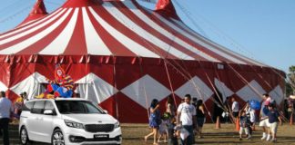 A white 2022 Kia Carnival is shown with a clown on its roof, parked outside of a circus tent.