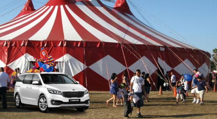 A white 2022 Kia Carnival is shown with a clown on its roof, parked outside of a circus tent.