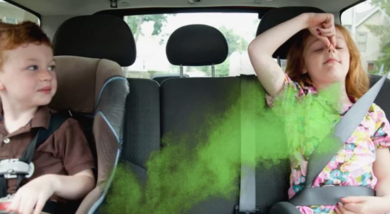 A close up shows a boy in a car seat farting while his sister is pinching her nose.