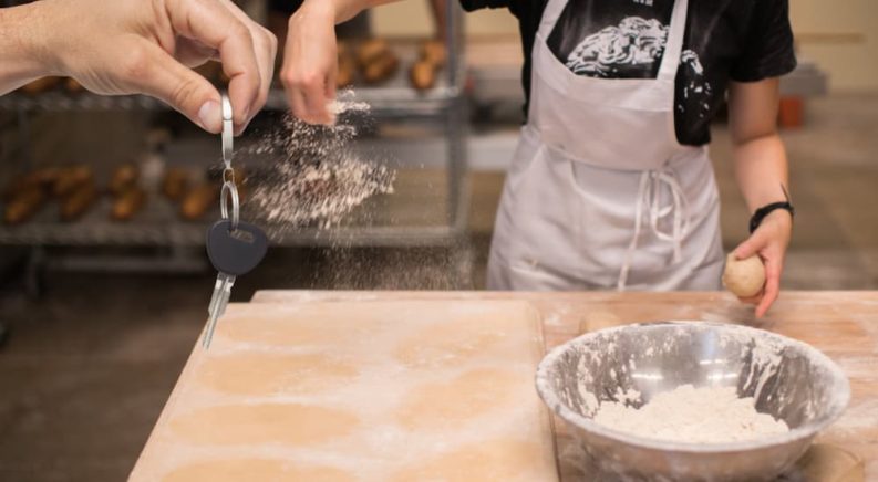A hand is shown holding a set of car keys in front of a person sprinkling flour on a table.