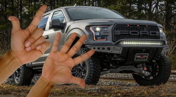 A hand holding up two fingers and a hand holing up five are shown in front of a dark grey 2021 Ford F-150 Raptor.