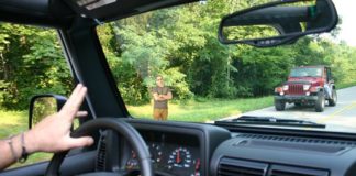 Two Jeep wranglers are shown passing each other while waving, driving past a man with his arms crossed.