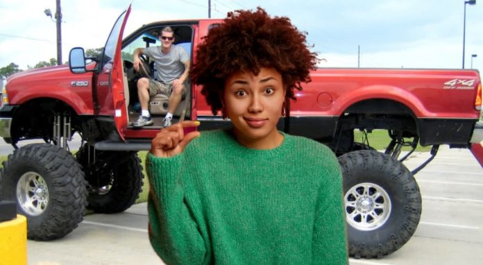 A woman is making a tiny penis reference with with fingers in front of a man in a lifted red truck.